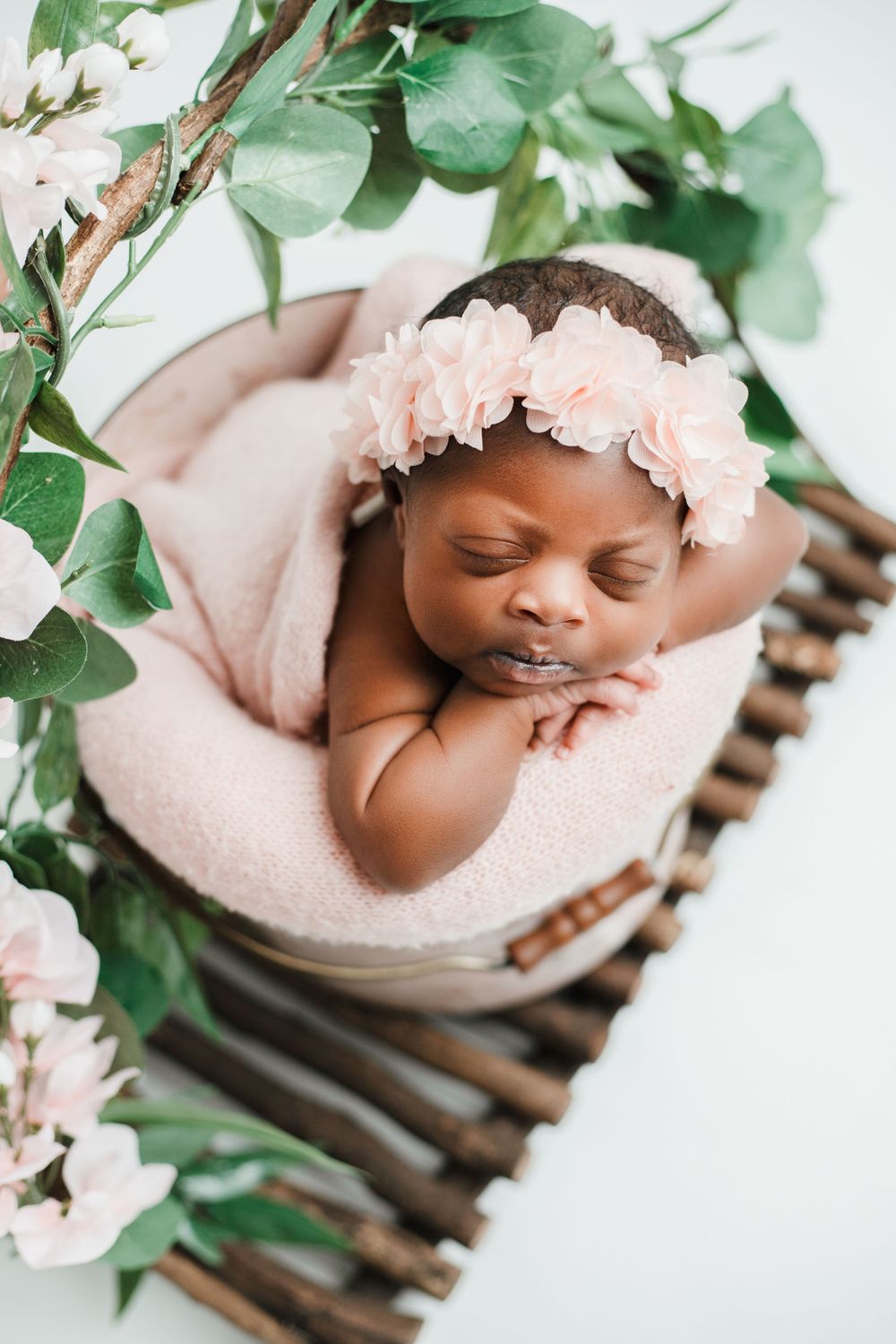 Peaceful sleeping newborn in pink headband nestled in blush blanket with green foliage decor.