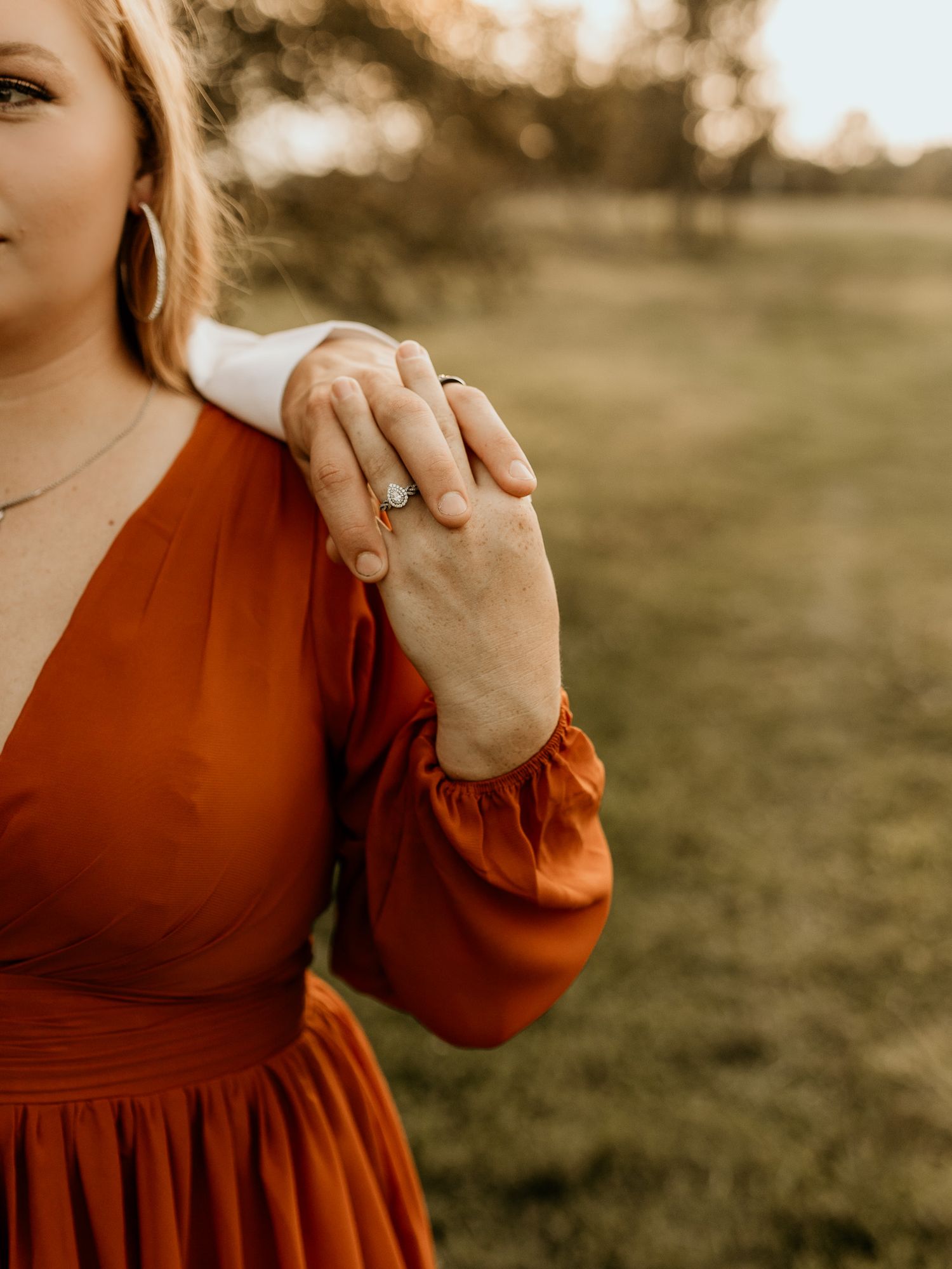 A close-up shot of hands clasped together with an engagement ring, wearing a rust-colored dress outdoors.