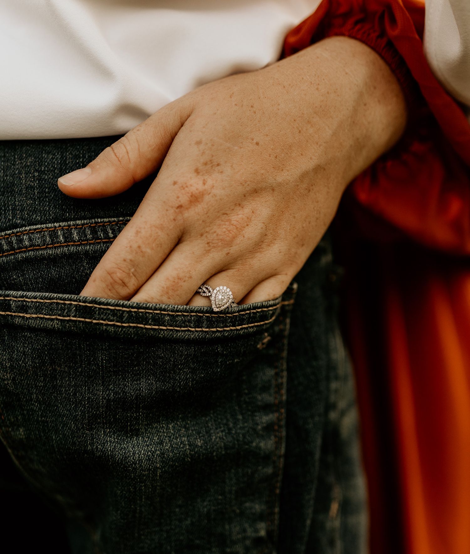 A hand wearing an engagement ring rests in a denim pocket while wearing a red sleeve.