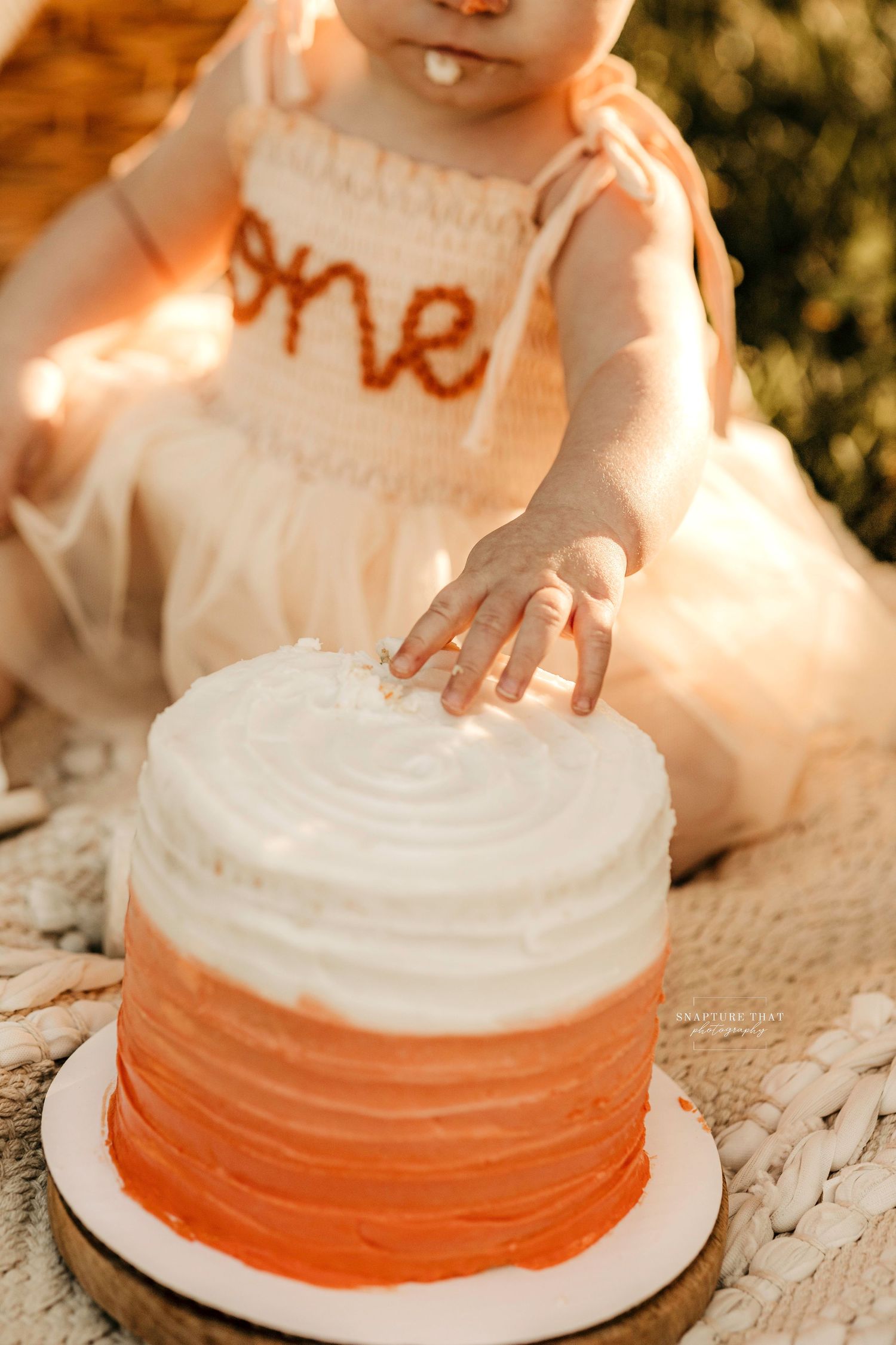Baby reaching for white and orange ombre birthday cake wearing dress with 'one' text.