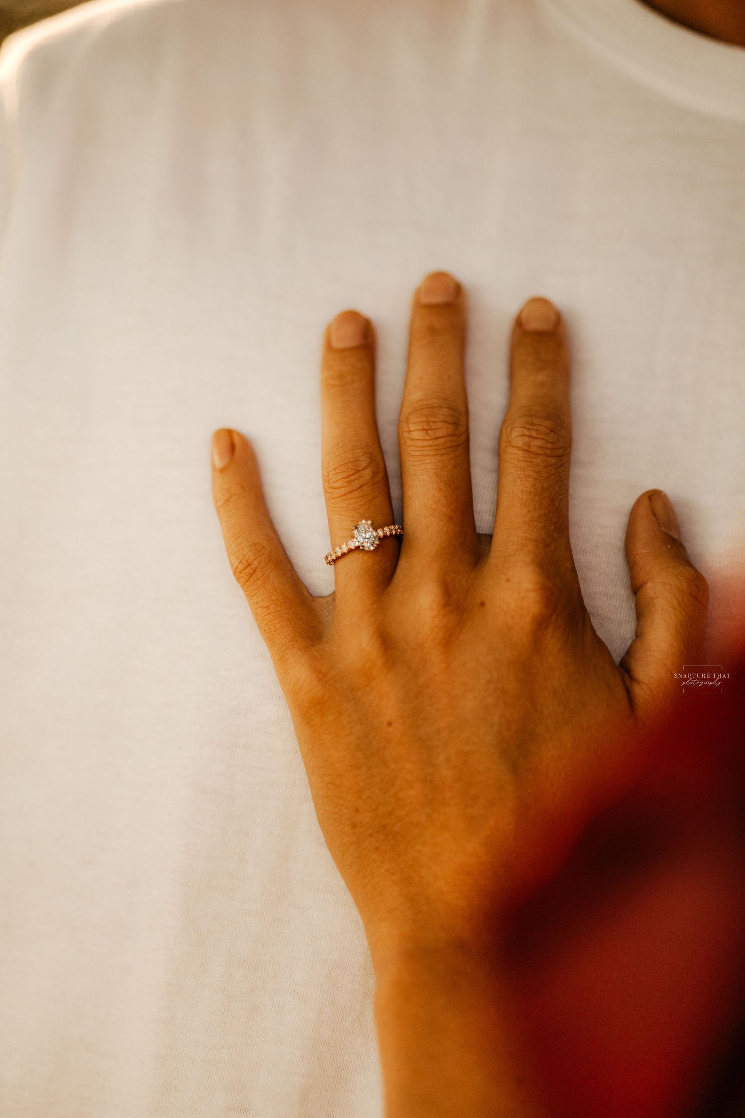 Hand with engagement ring resting against white fabric in soft lighting.