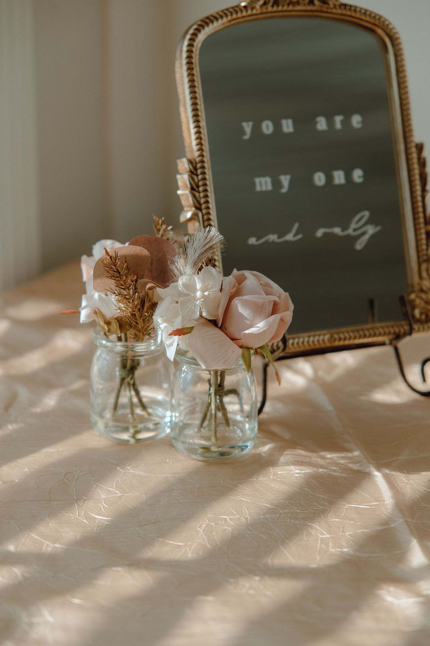 Delicate pink roses in glass vases beside vintage gold-framed mirror with text.