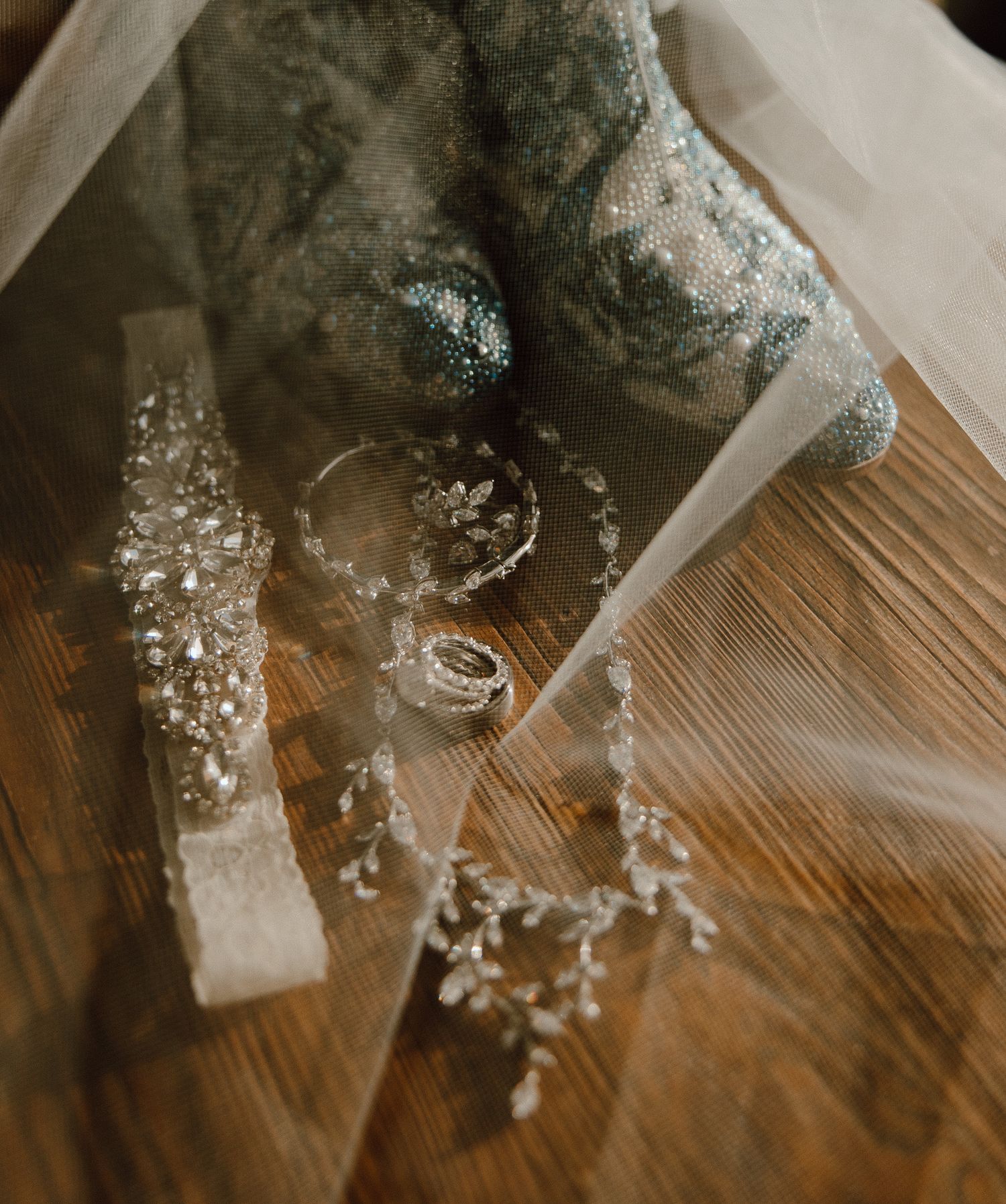 Wedding rings and jewelry arranged with patterned boot and veil on wooden surface.