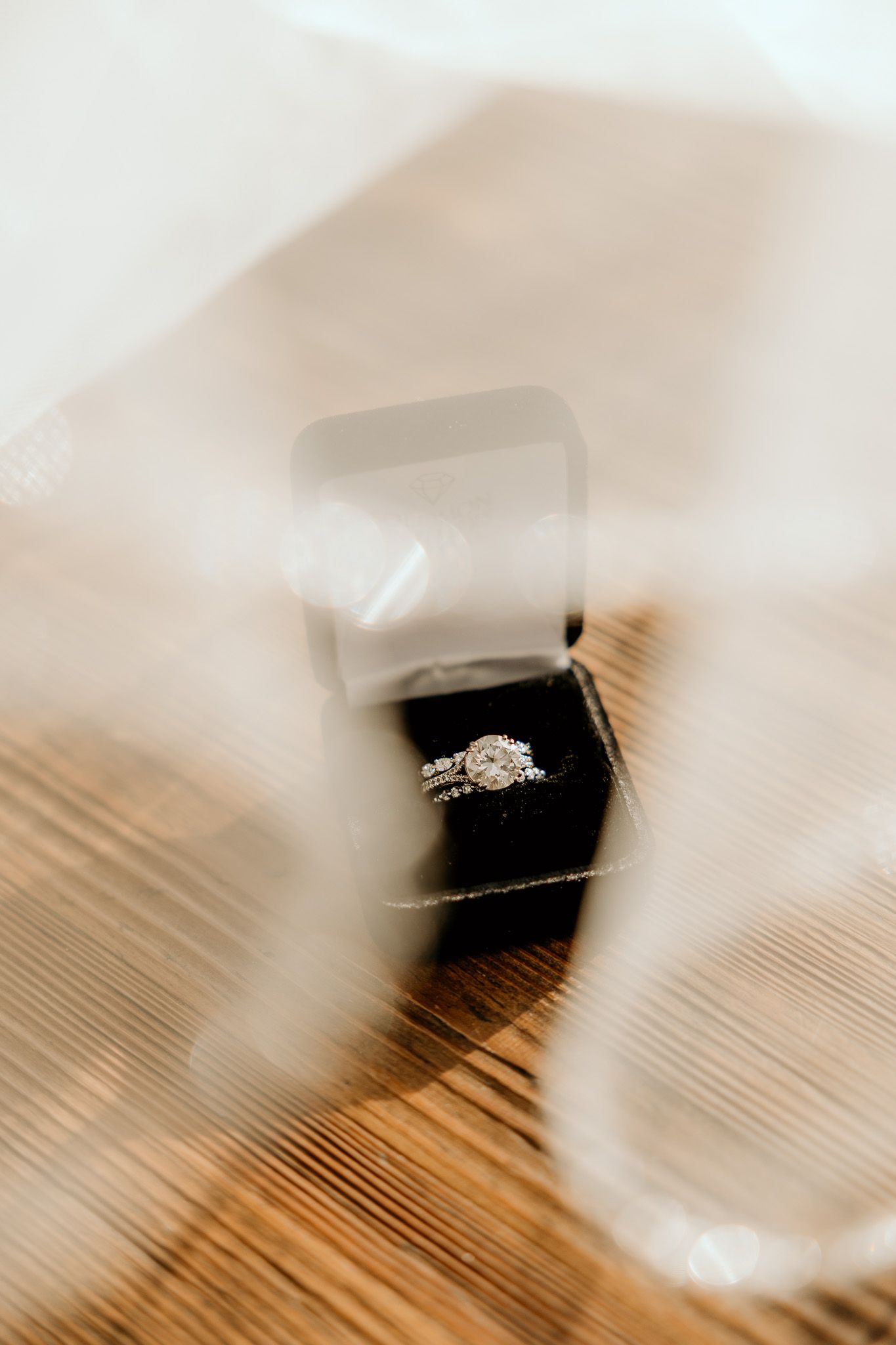 Elegant diamond engagement ring in black box photographed through sheer white fabric on wooden surface.
