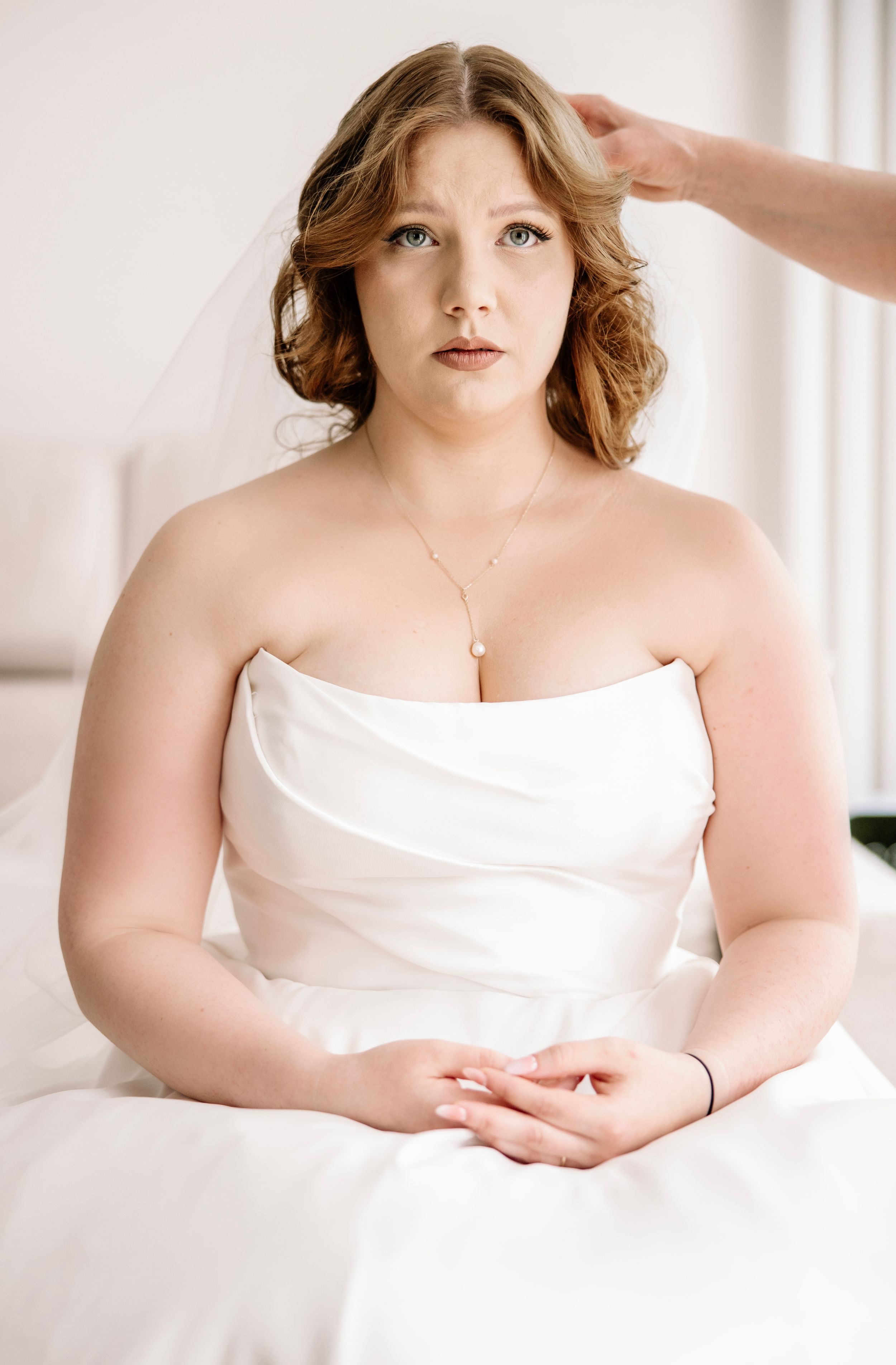 Plus-size bride in strapless white wedding gown with veil, pearl necklace, getting hair styled before ceremony.