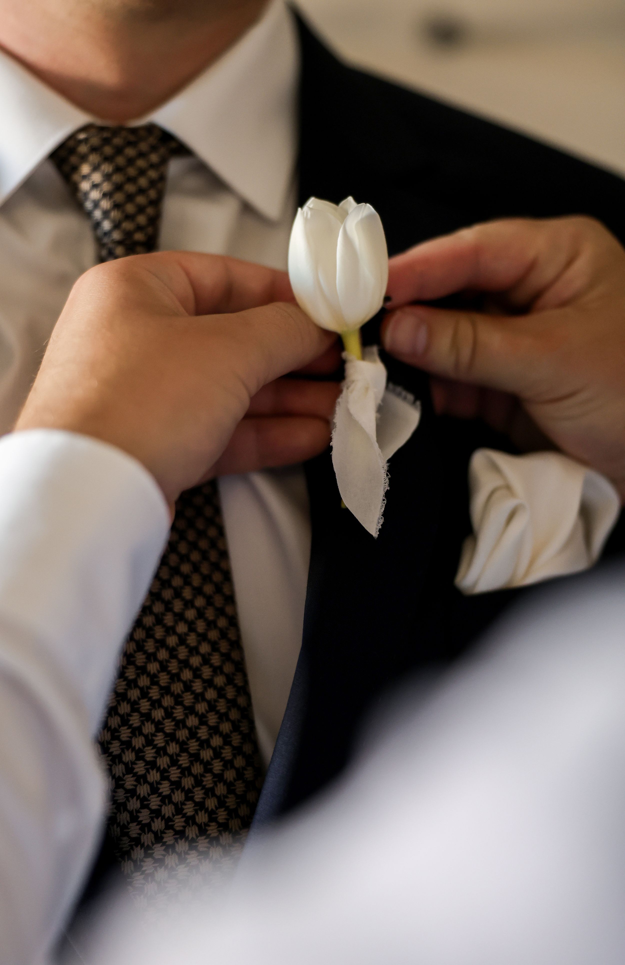 Hands pinning a white tulip boutonniere onto a groom's dark suit lapel at a wedding ceremony.