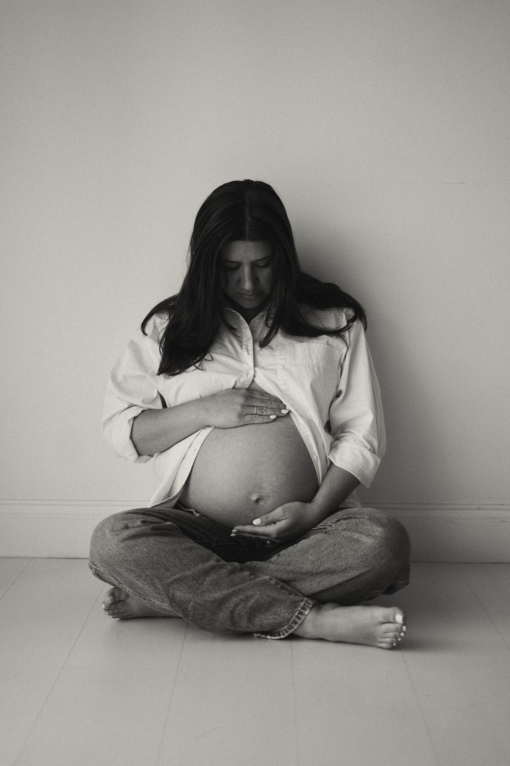 Black and white photo of a pregnant woman sitting cross-legged on the floor cradling her baby bump.