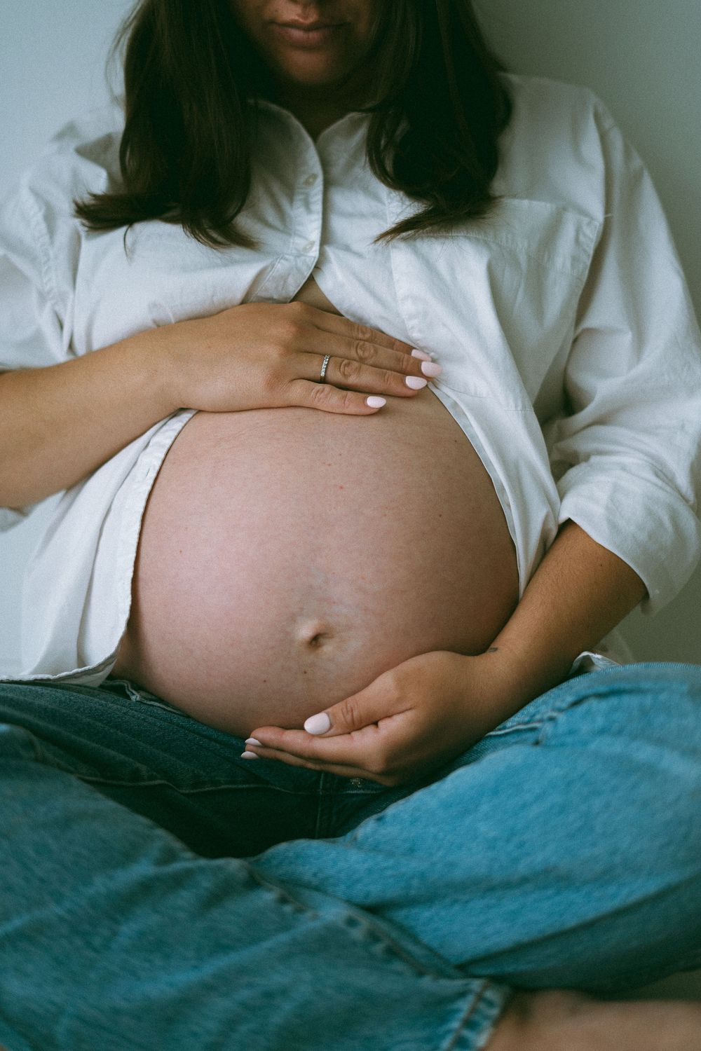 Close-up of a pregnant womans bare belly held gently in her hands wearing an open white shirt and jeans.