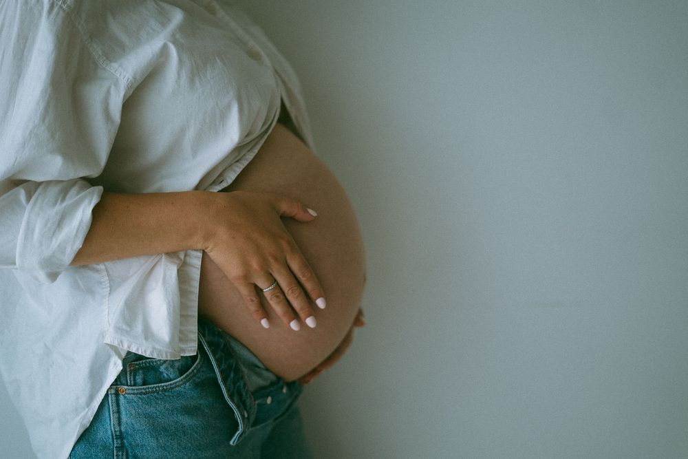Pregnant woman in white shirt and jeans cradling her bare baby bump against a neutral background.