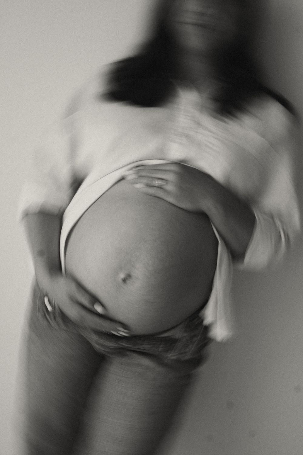 Black and white blurred artistic photo of pregnant woman holding her bare belly in white shirt and jeans.
