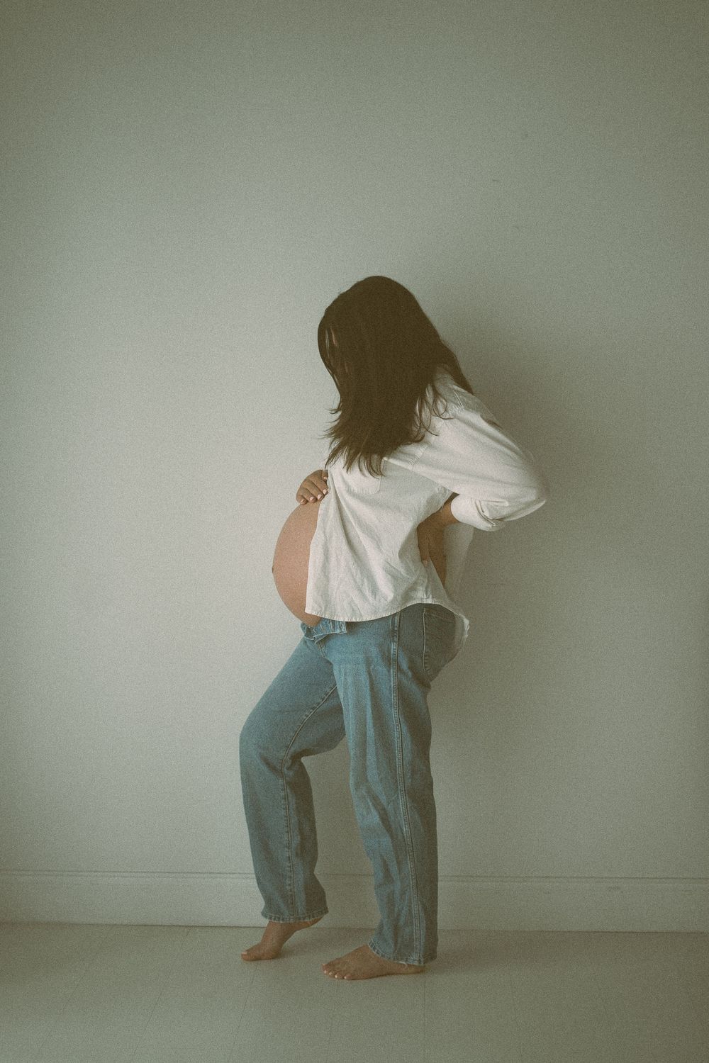 Pregnant woman in white shirt and jeans standing barefoot against a light wall showing her baby bump.