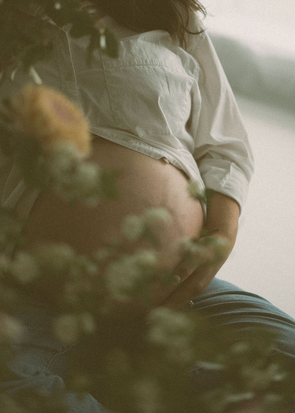 Pregnant woman in white shirt and jeans sitting outdoors with soft floral bokeh framing her baby bump.