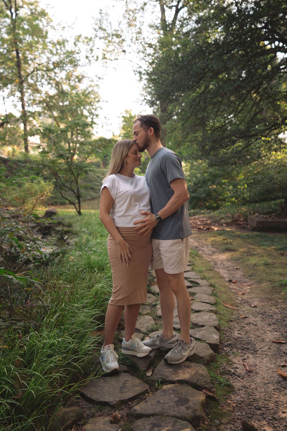 Expecting couple stands on a stone path in a lush green park, embracing and touching the pregnant belly.