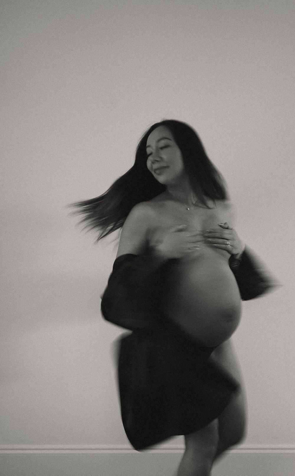 Black and white photo of a pregnant woman cradling her belly, smiling with hair flowing in motion.
