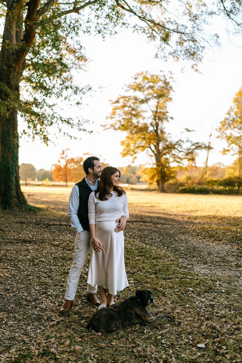 Expecting couple with dog standing under large oak tree during golden autumn sunset.