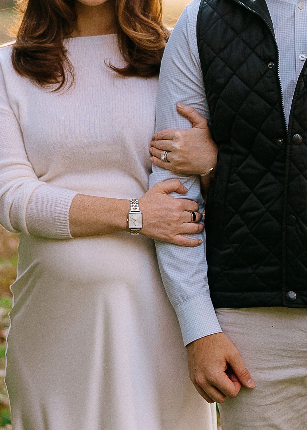 Pregnant woman in cream dress holds partner's arm outdoors, wearing a silver watch and wedding ring.