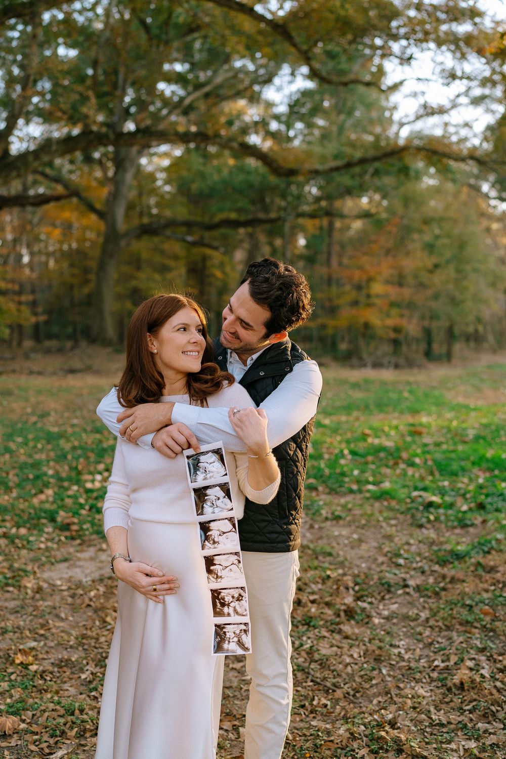 Couple in autumn park holding ultrasound photos, man embracing pregnant woman in white dress.
