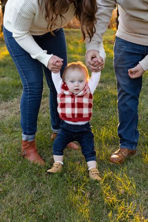 Parents helping small child in plaid vest take first steps on grassy lawn. Family session in Lake Geneva, Wisconsin.