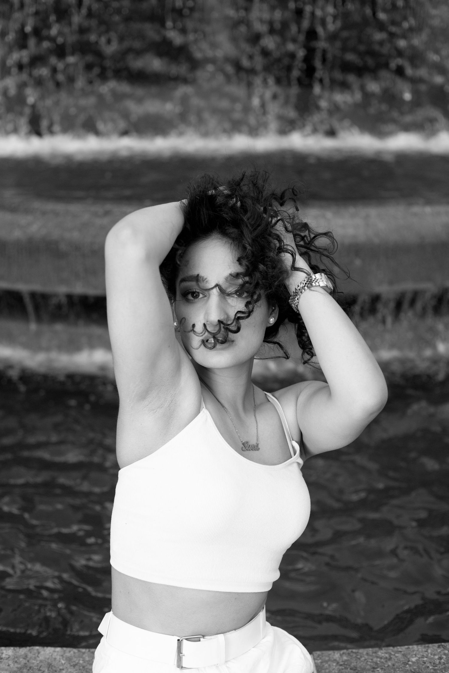 Black and white portrait of woman in white top posing with windswept hair near water.