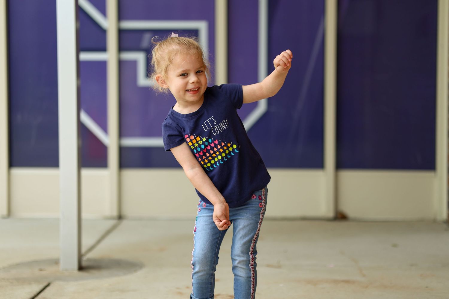 Child strikes playful dance pose against purple wall background.