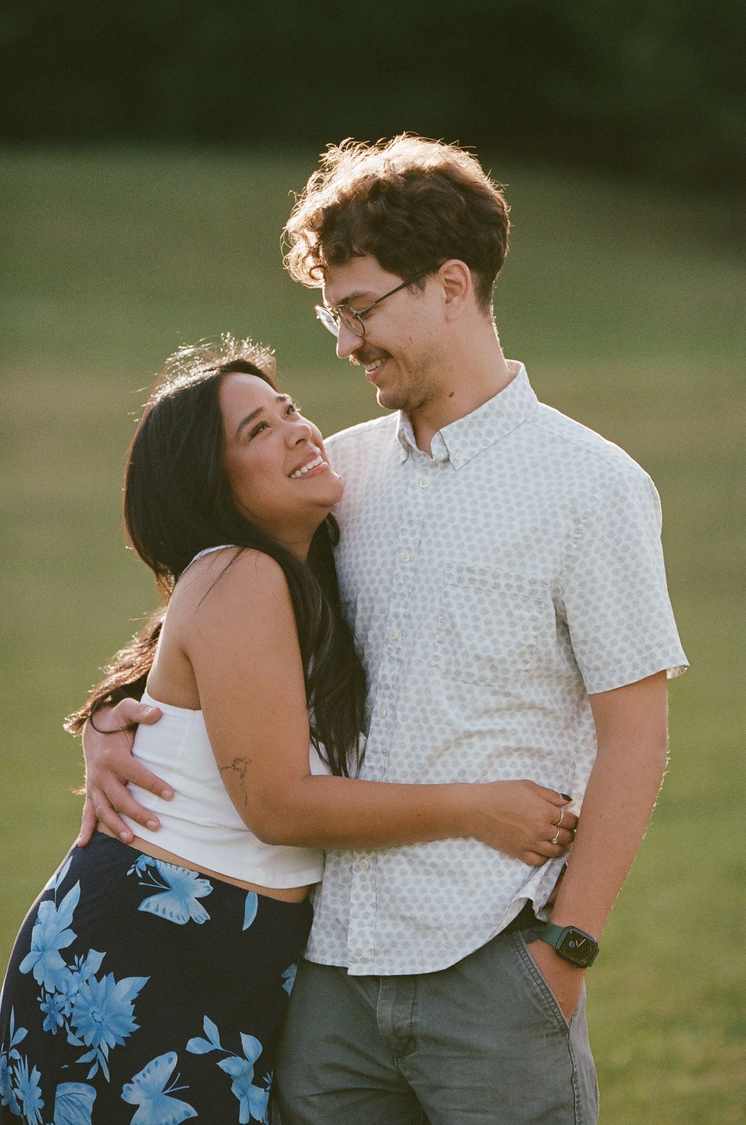 A couple laughs together during their engagement photos in a sunlit park setting.