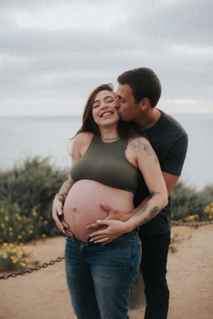 Joyful maternity photos of couple laughing and embracing outdoors with ocean view.