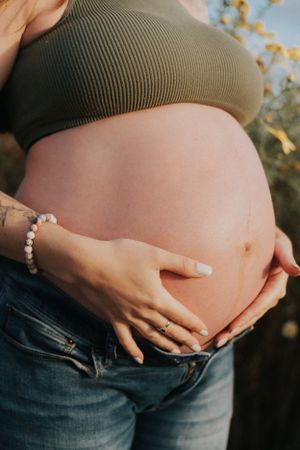 Close-up of hands cradling pregnant belly in olive green top against sunset light.