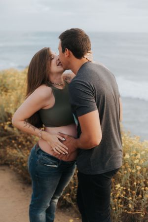 Romantic maternity photoshoot with couple embracing on coastal cliffs with yellow wildflowers.