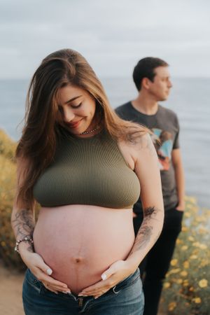Pregnant woman in olive green top shows her tattooed belly against ocean backdrop.