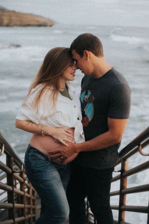 Couple shares intimate moment during sunset maternity shoot on coastal boardwalk.