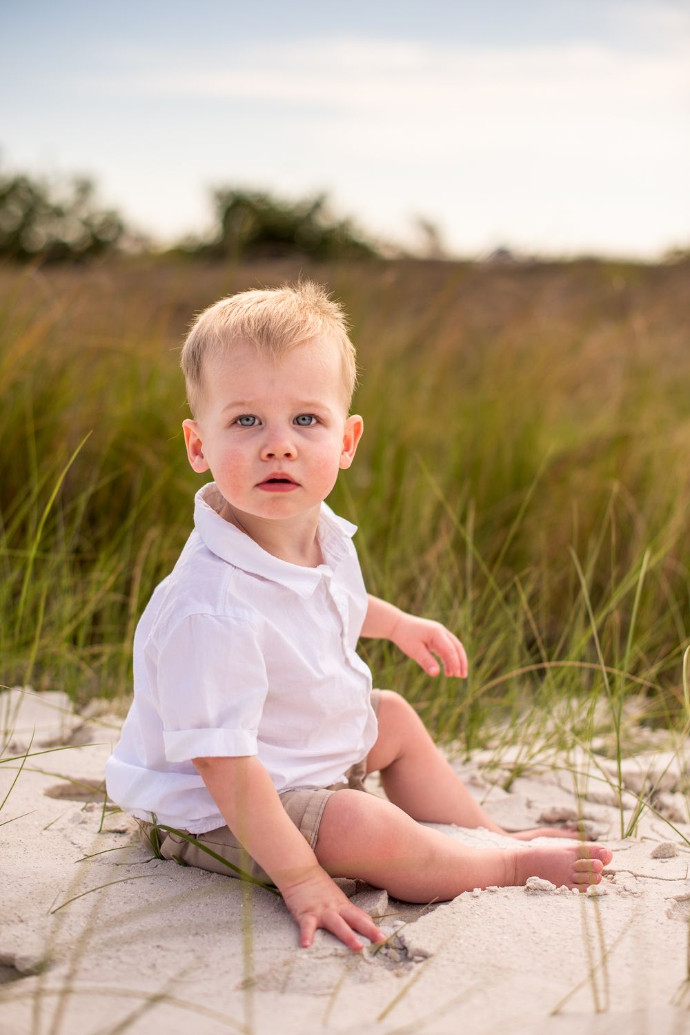 Child in white shirt sits on sandy beach during golden hour photography session.