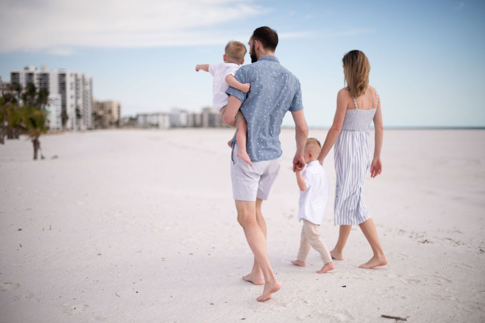 Family walking barefoot together along pristine white sand beach with buildings in background.