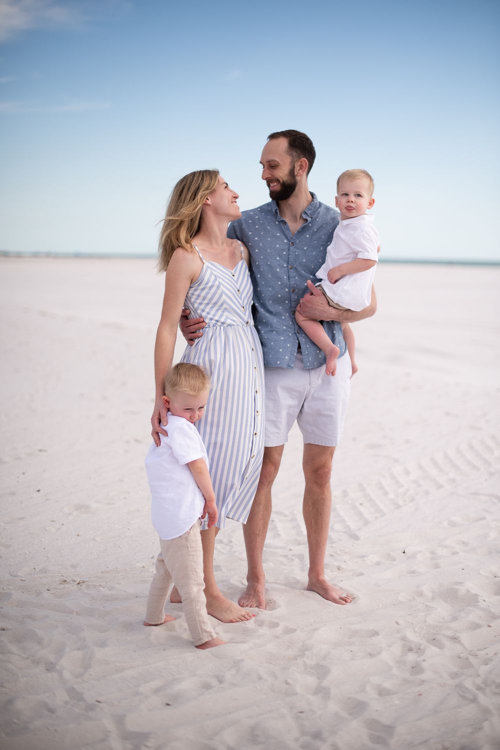 Family of four wearing coordinated beach outfits poses together on white sandy shoreline.