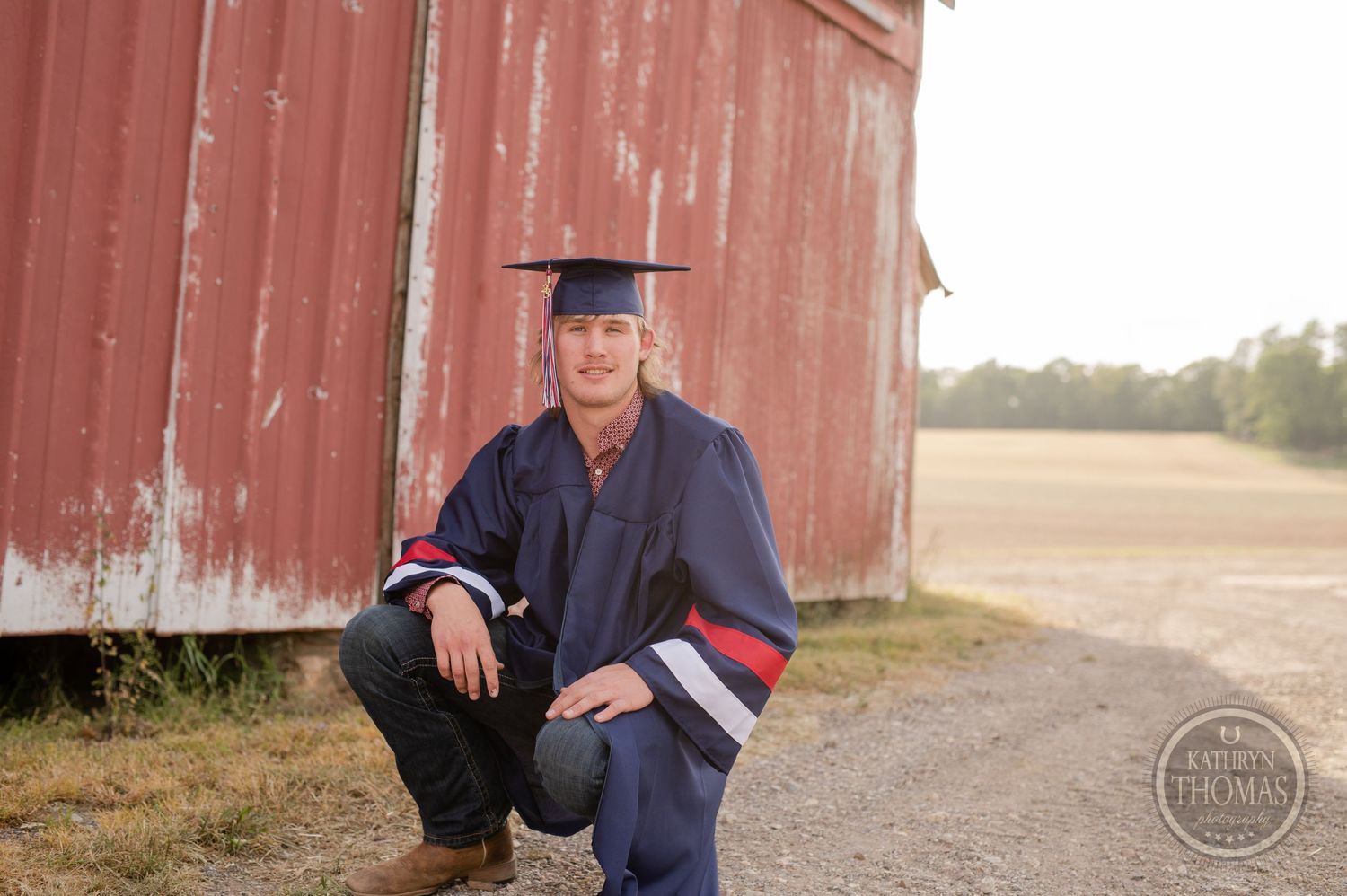 Graduate in academic regalia crouches for a photo by rustic barn.