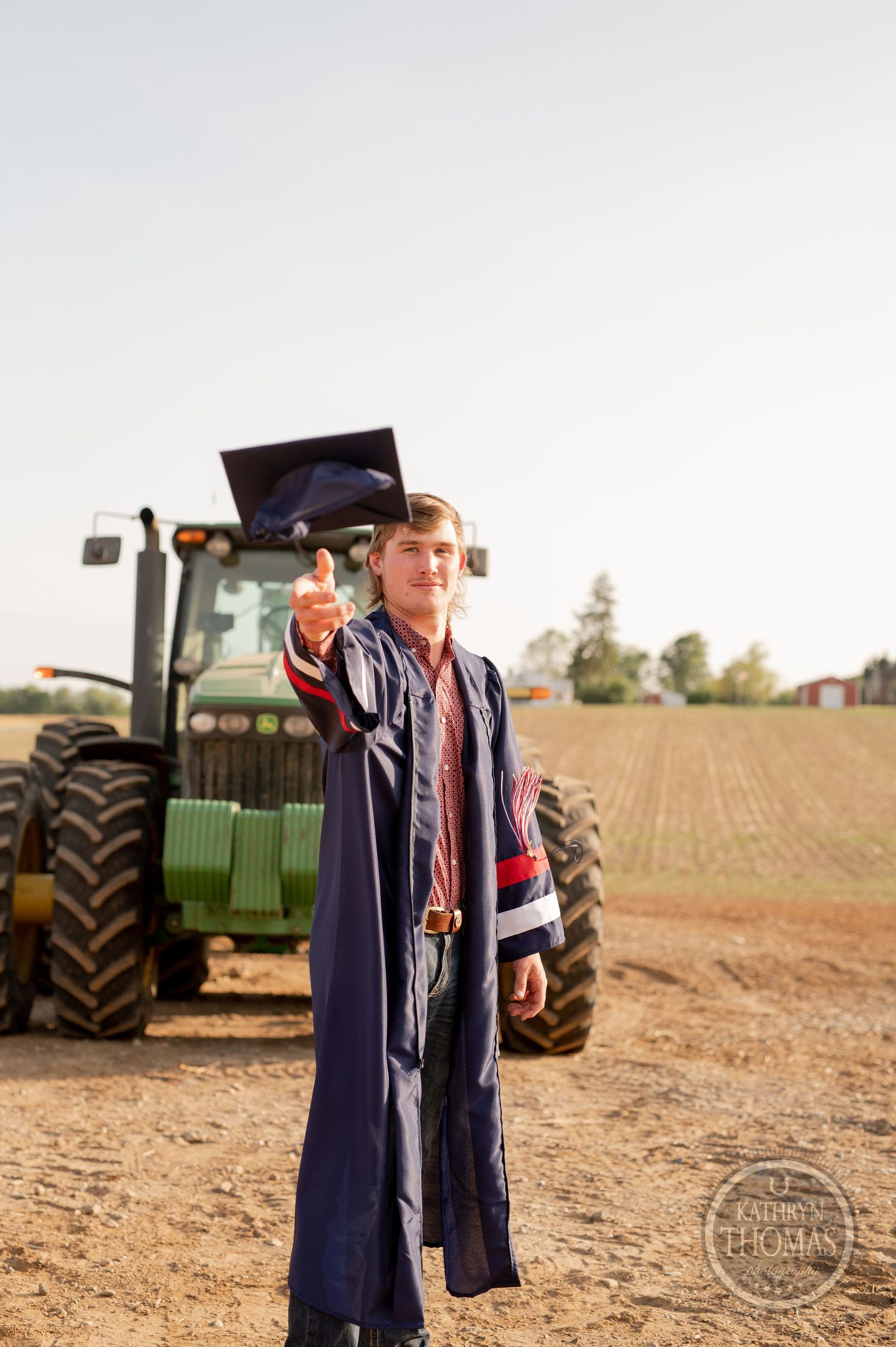 Graduate reaches for falling mortarboard in front of John Deere tractor.