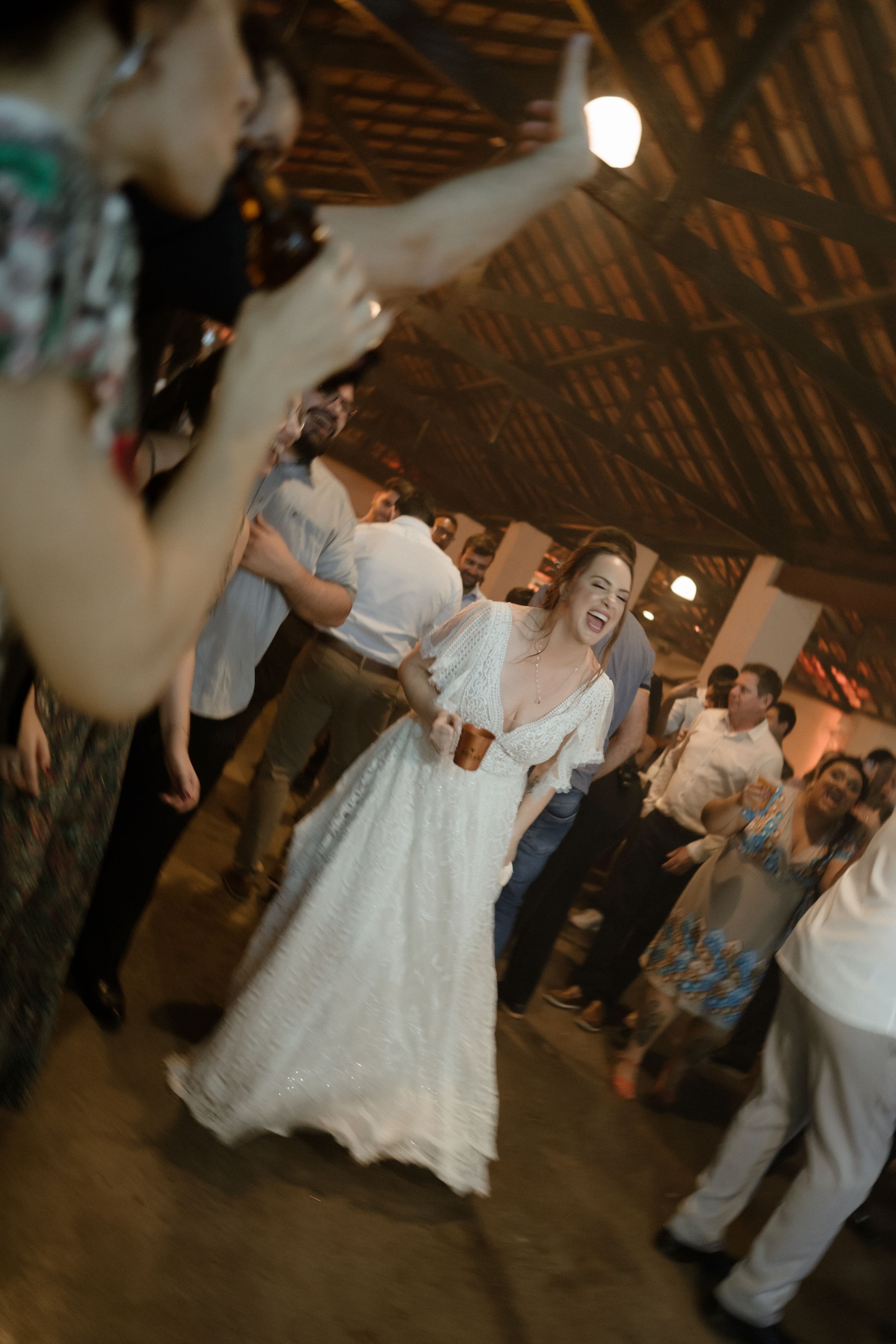A bride in a white lace dress dances joyfully at her wedding reception under rustic wooden beams.