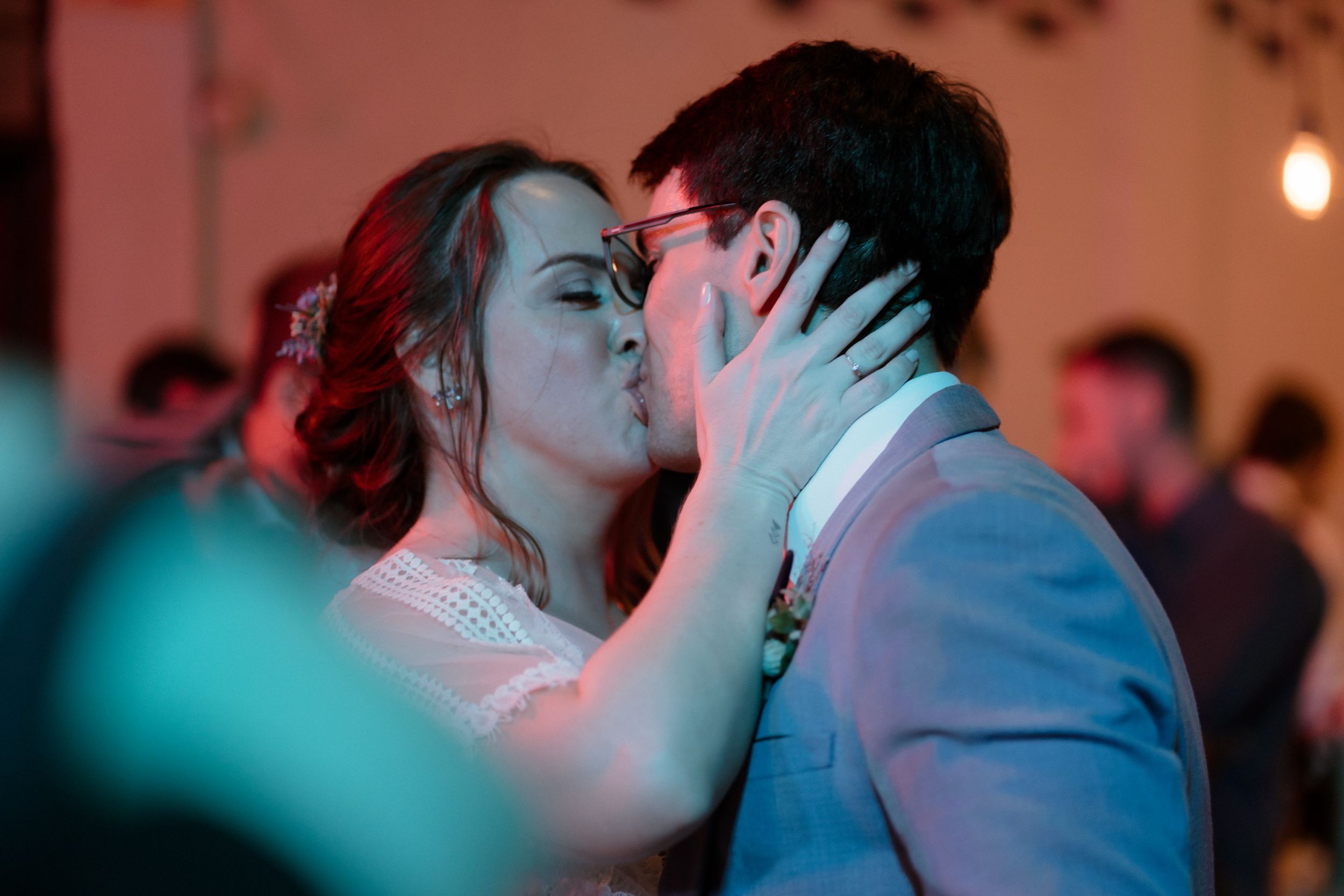 A romantic kiss shared between a couple during a wedding reception with atmospheric teal lighting.