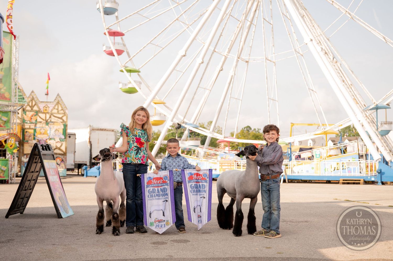 Show animals and handlers stand with award banners in front of large ferris wheel at county fair.