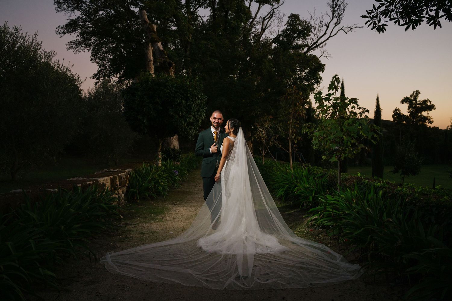A couple embraces on a garden path at dusk with a dramatic flowing cathedral-length veil trailing behind them - WeddingInvestment