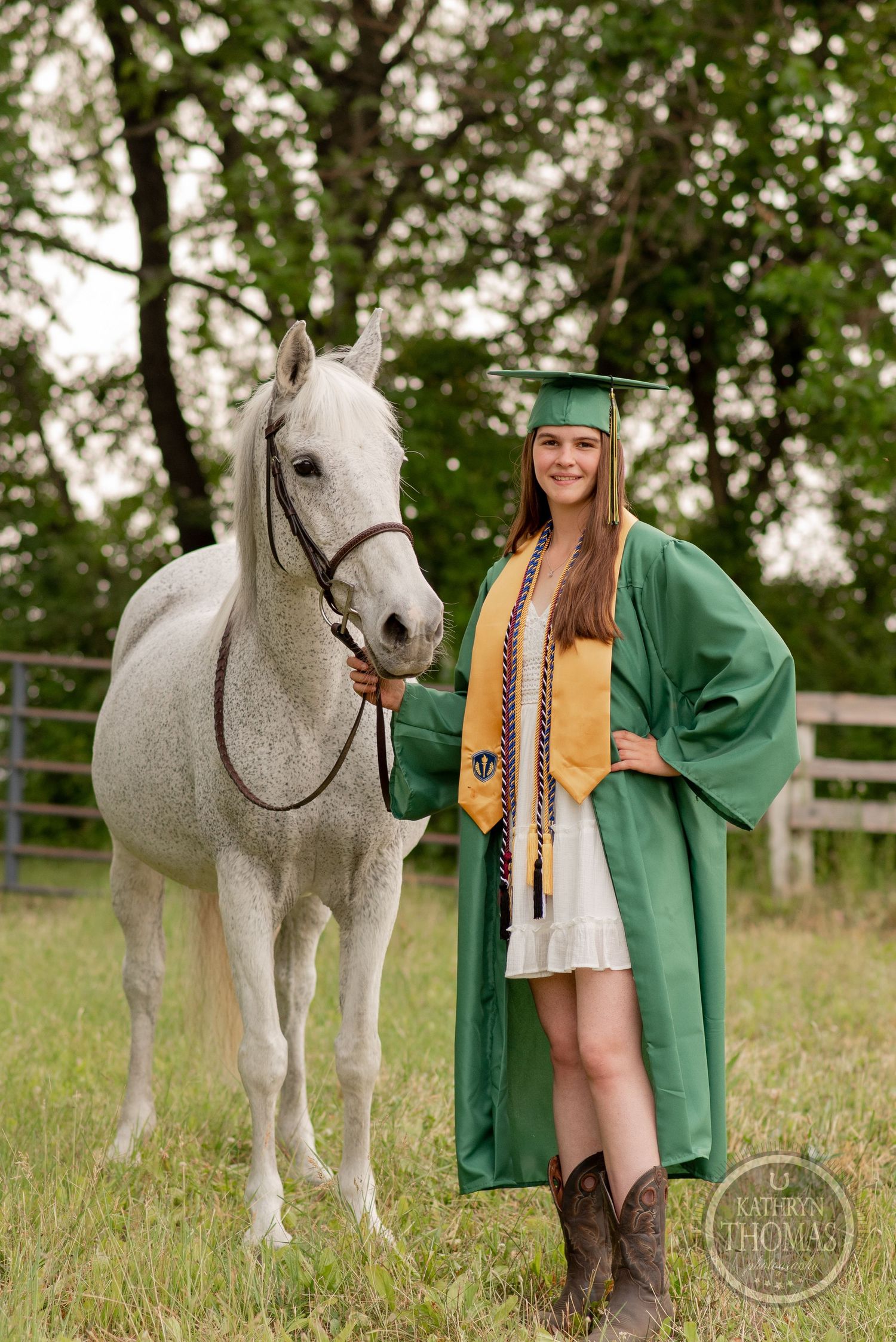 Graduate in green cap and gown poses with elegant white horse by wooden fence.