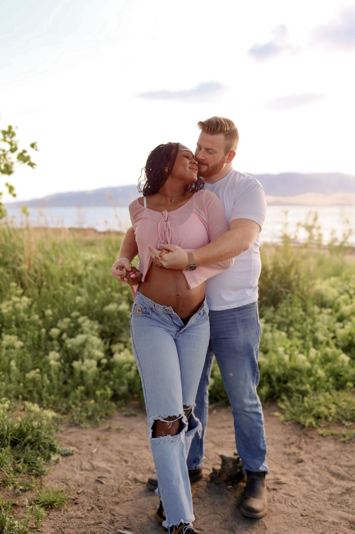 Couple shares a romantic embrace in a field of wildflowers near a lake at sunset.