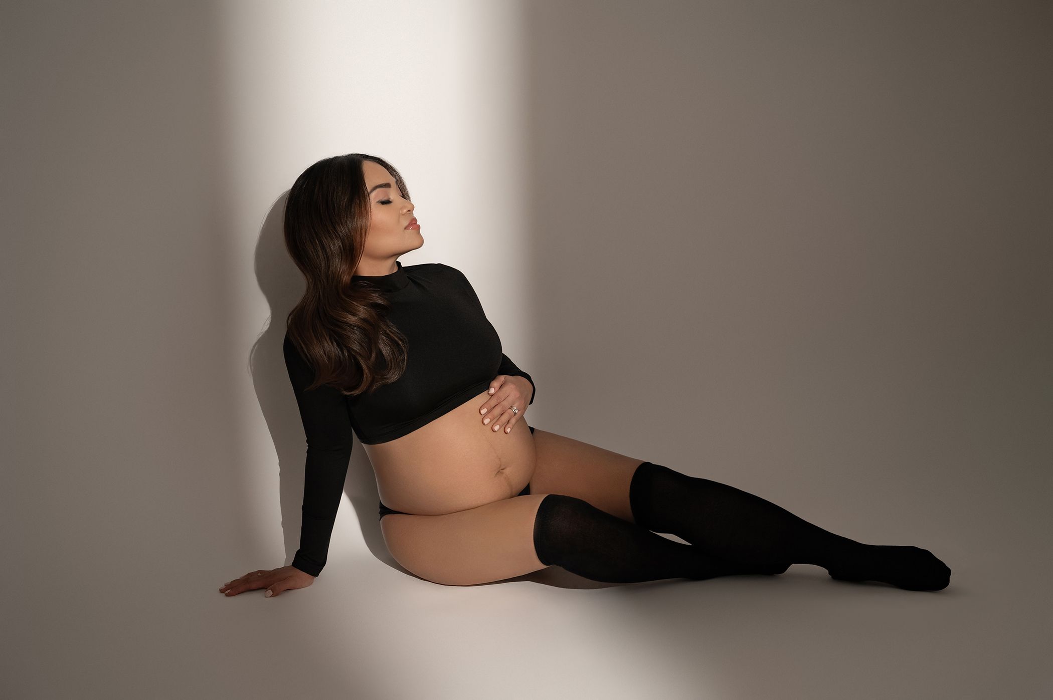 Pregnant woman in black crop top and thigh-high socks posing against a white studio wall.