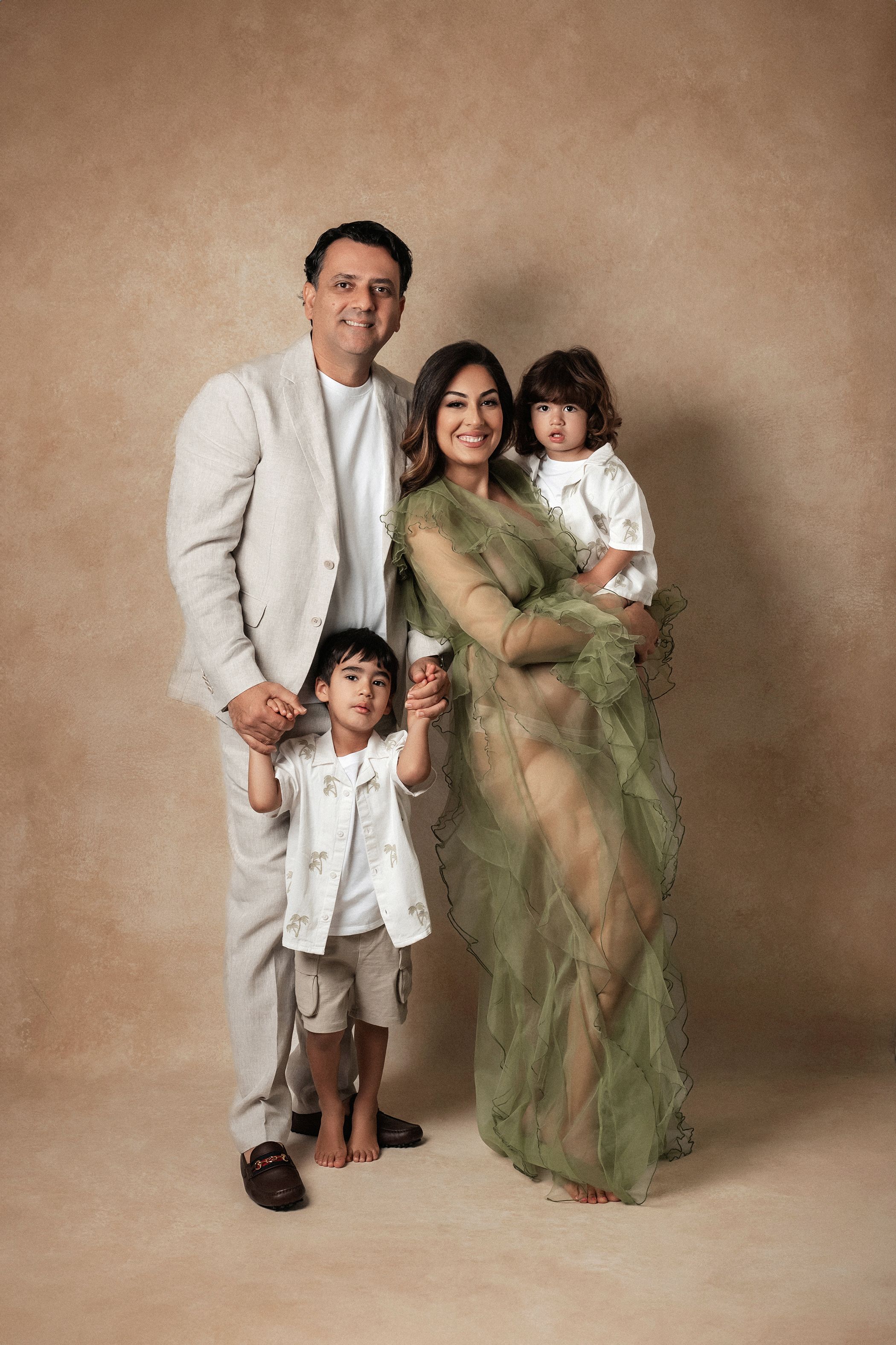 A happy family of four poses together, mother in green tulle maternity gown, on a beige backdrop.
