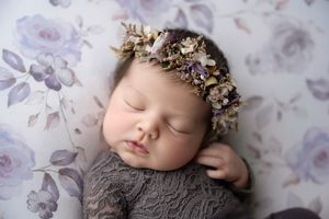 Serene newborn baby in gray lace outfit wearing purple dried floral crown on lavender floral backdrop.