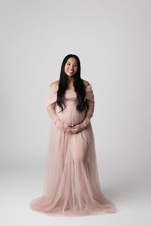 A smiling pregnant Asian woman in an elegant off-shoulder blush pink tulle maternity gown.