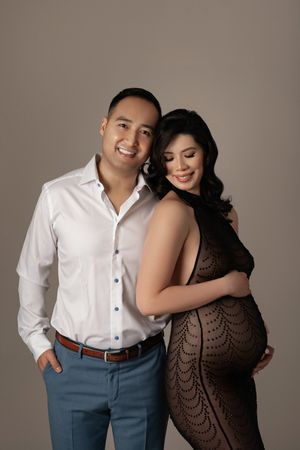 Smiling couple in maternity photoshoot, man in white shirt and woman in black lace dress cradling baby bump.
