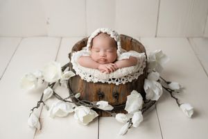 Sleeping newborn baby in white lace bonnet posed in rustic wooden bucket surrounded by white roses.