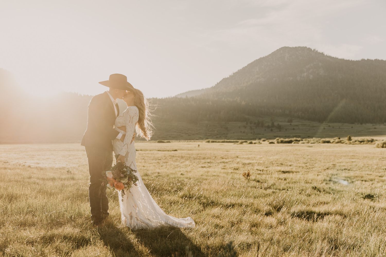 A couple stands in a golden meadow during sunset with mountains in the background.