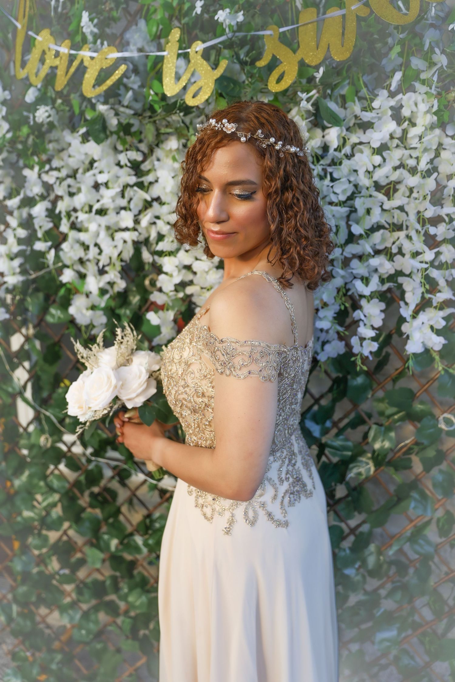 Bride in beaded off-shoulder gown poses with white flowers against floral backdrop and gold banner.