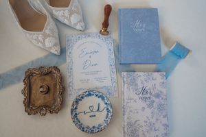 Elegant wedding flat lay with blue vow books, floral invitation, lace heels, wax seal, and vintage ring dish in soft blue tones.
